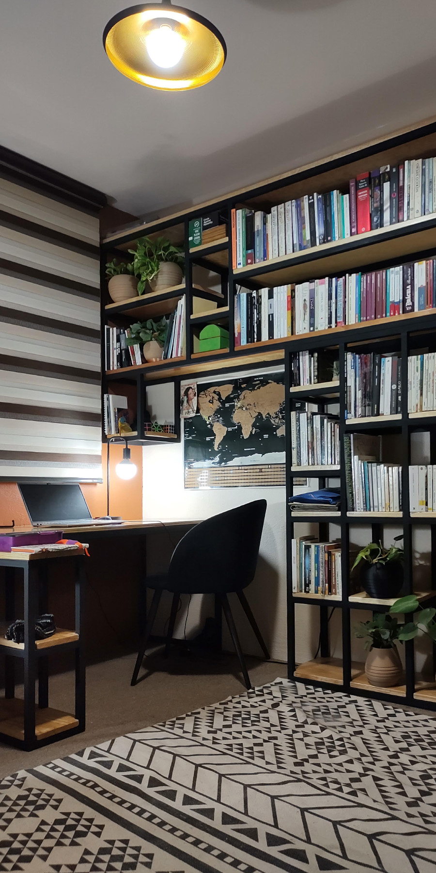 Sala de trabajo con estantes metálicos negros de base de madera adornados con plantas y libros, una cortina rayada adorna la ventana de la sala.