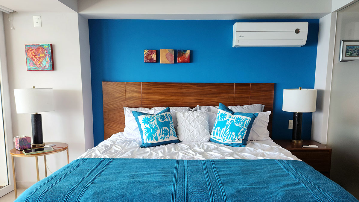 Cuarto de color azul celeste con una gran ventana que da a un balcón. Está adornado con una cama con cabecera de madera color cedro, una cama king size con cobijas y almohadas azules y blancas. También cuenta con aire acondicionado.