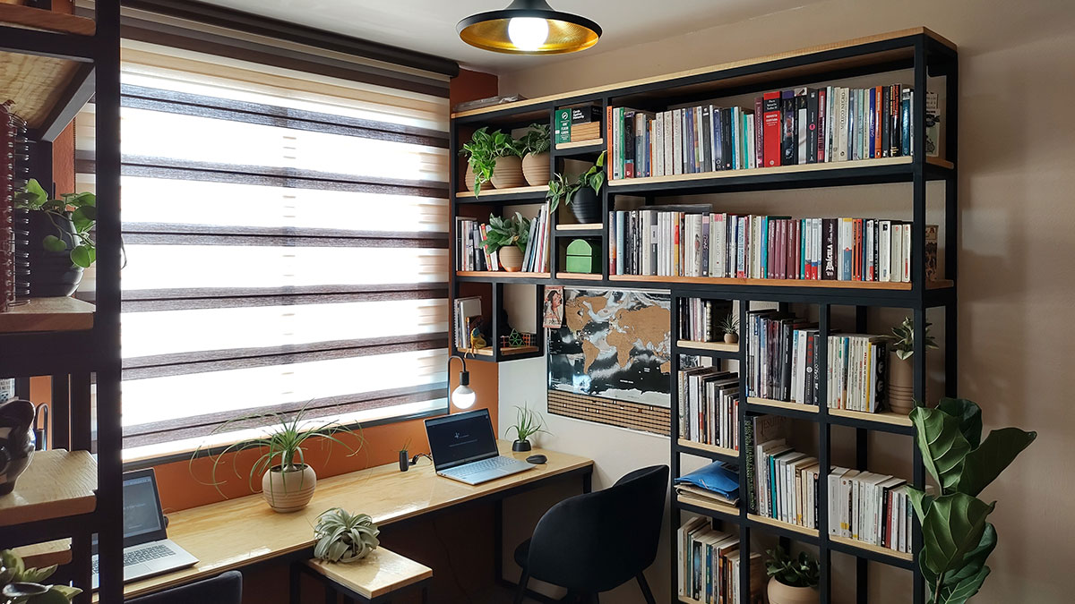 Sala de trabajo con estantes metálicos negros y madera, adornados con plantas y libros en cada muro lateral. Una mesa de trabajo de madera con espacio para dos personas, cerca de una ventana con una cortina a rayas.