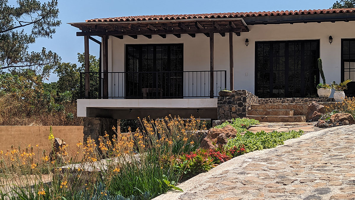 Casa de muros blancos con techo de tejas y un balcón. La fachada tiene ventanas altas y una puerta de madera. La entrada y el camino están empedrados y adornados con diversas flores y macetas con cactus.