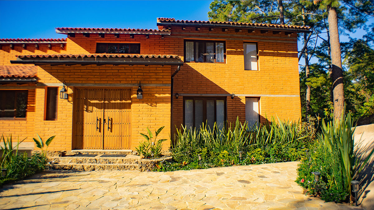 Casa de ladrillo naranja con techo de tejas y una puerta de madera. La fachada cuenta con múltiples ventanas rectangulares y lámparas de metal oscuro. El camino está empedrado y aplanado, y está adornado con plantas.