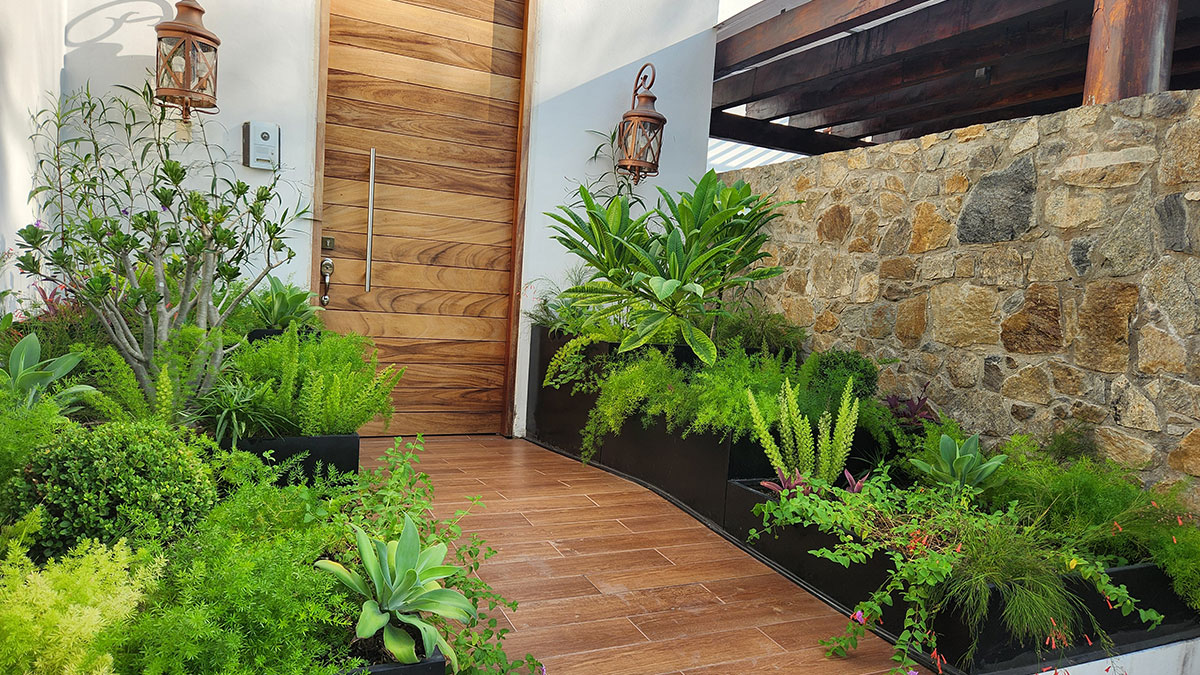 Casa de ladrillo naranja con techo de tejas y una puerta de madera. La fachada cuenta con múltiples ventanas rectangulares y lámparas de metal oscuro. El camino está empedrado y aplanado, y está adornado con plantas.